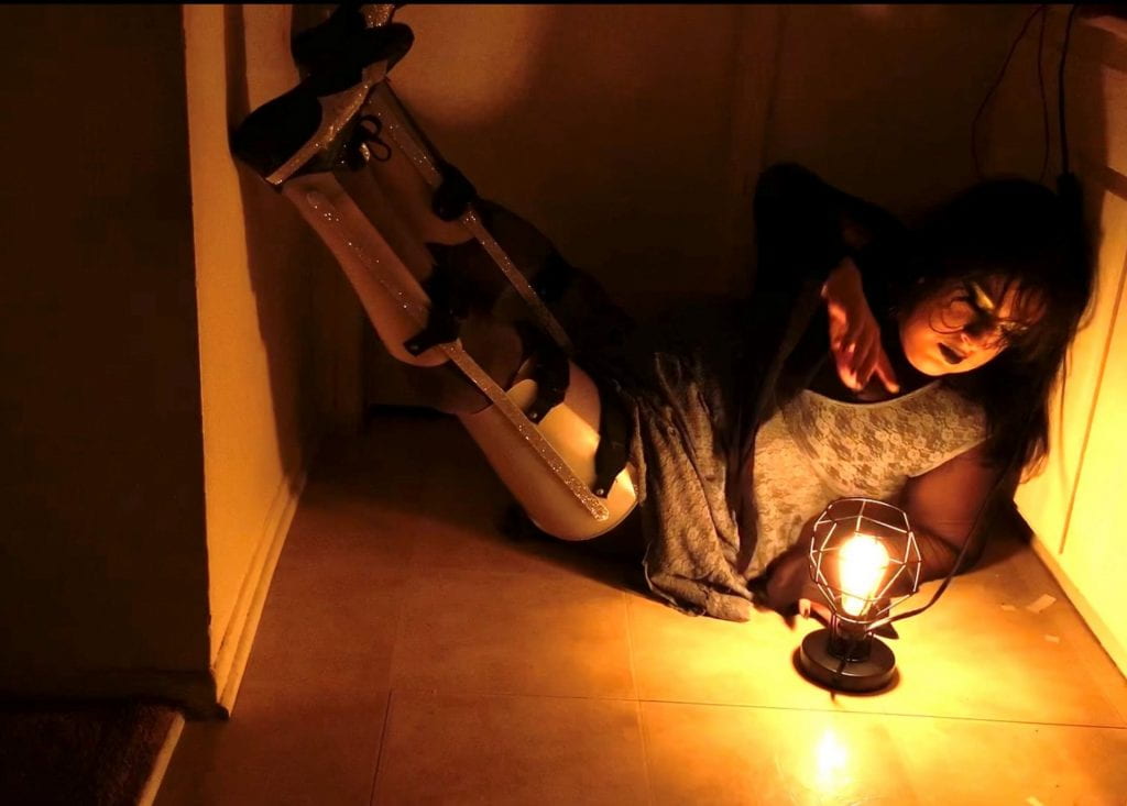 A photo of Vanessa in a dark lit room. She is lying on the pale yellow tiled floor with her sparkly braced legs on the wall. She is wearing a gray long lace dress with a mesh long sleeve shirt underneath. Her makeup is channeling Goth aesthetics and she is gazing down at an industrial lamp.
